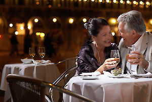 A couple is sitting at a table in a restaurant, holding wine glasses and looking at each other.