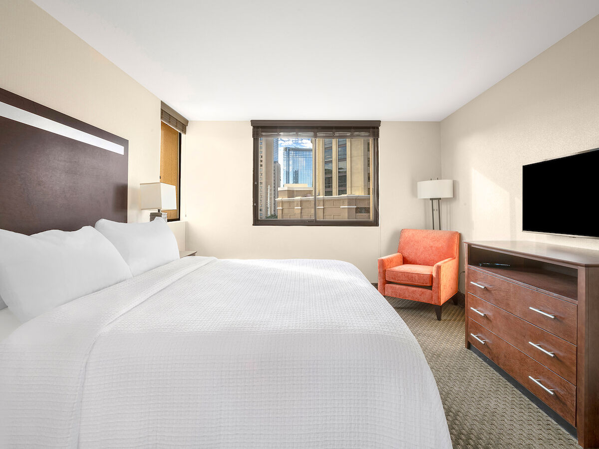 A bright hotel room with a large bed, TV on a dresser, an orange chair, lamp, and window with a city view in the background.
