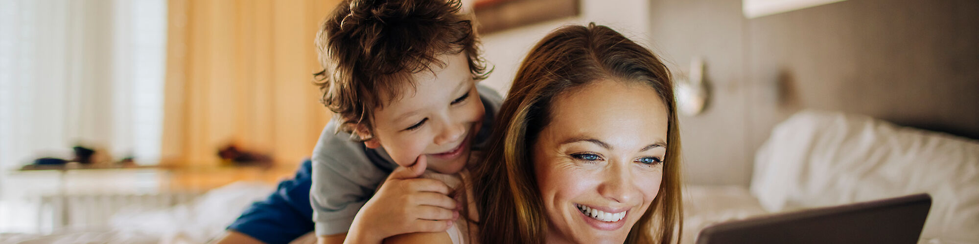 A woman is lying on a bed, using a tablet, while a child is happily climbing on her back, both smiling and enjoying their time together.