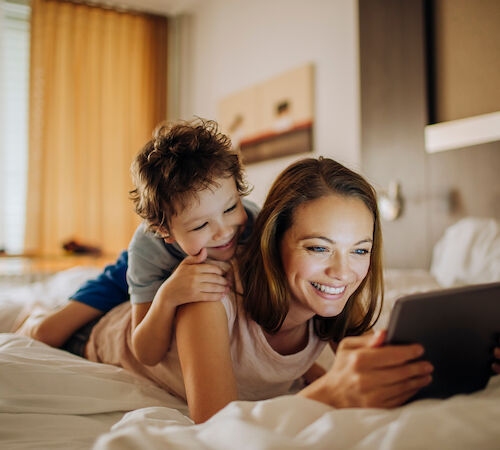 A woman and a boy are lying on a bed, looking at a tablet.