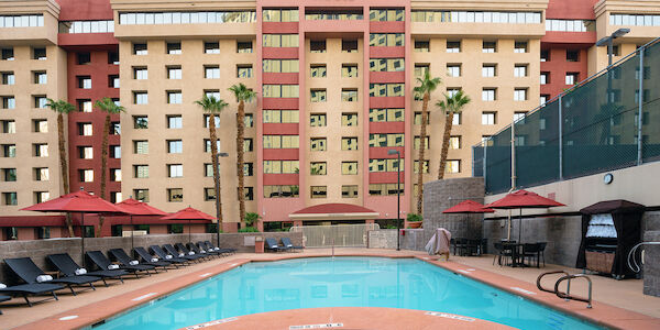 A hotel pool area with lounge chairs, umbrellas, and palm trees in front of a large hotel building.