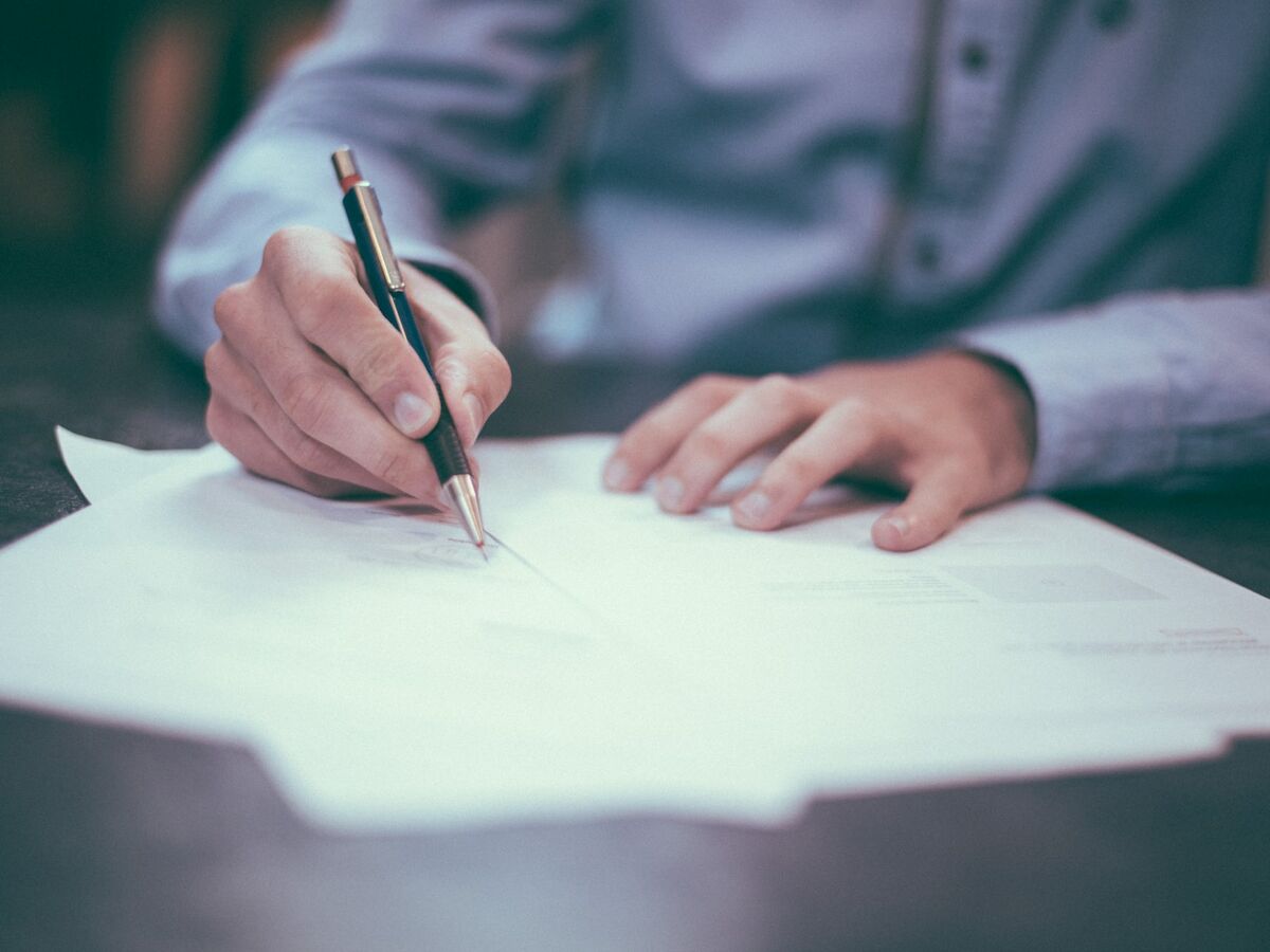 A person is writing on documents at a desk, using a pen and surrounded by paper. The focus is on the hand holding the pen.