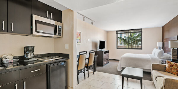 A wide view of a hotel suite with a kitchenette, dining area, sofa, bed, and TV.