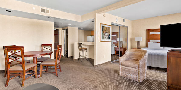 A wide view of a hotel suite with a kitchenette, dining table, sofa, bed, and TV.