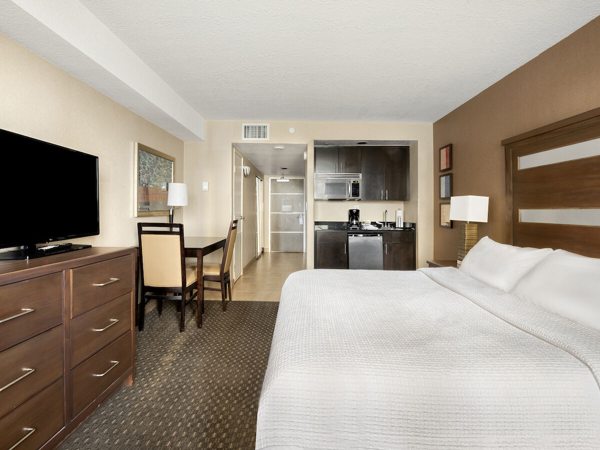 A hotel room with a large bed, TV on a dresser, small dining area, and a kitchenette with a microwave and fridge in the background.