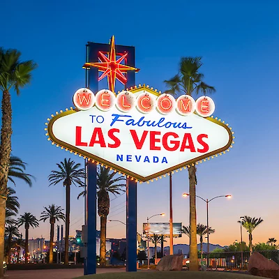 The image shows the iconic "Welcome to Fabulous Las Vegas, Nevada" sign at dusk, surrounded by palm trees, with the cityscape in the background.