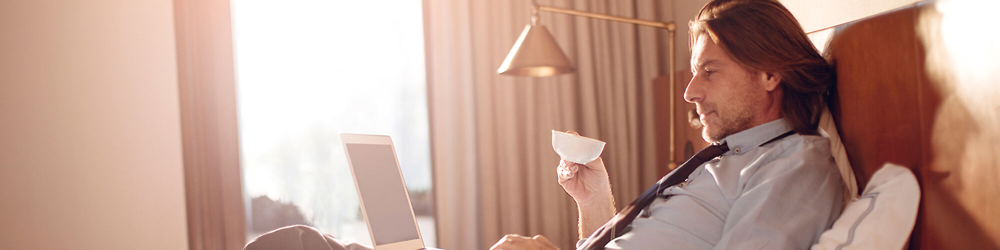 A man is lounging on a bed, using a laptop, and holding a cup, possibly in a hotel room with natural light streaming in from the window.