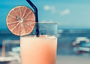 A refreshing beverage with a lime slice garnish and a straw is held against a sunny beach scene in the background.