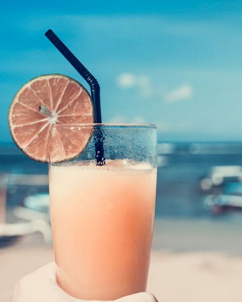 A refreshing beverage with a lime slice garnish and a straw is held against a sunny beach scene in the background.
