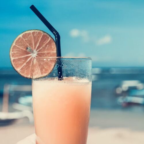 A refreshing beverage with a lime slice garnish and a straw is held against a sunny beach scene in the background.