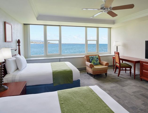 A hotel room with two beds, a desk, a chair, and a TV, with a large window showing an ocean view.